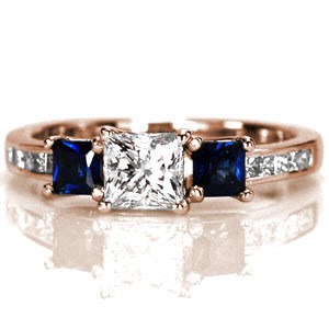 Tricia is a eye-catching princess cut three stone design. Two colorful blue sapphires are positioned on either side of a 0.50 carat square brilliant. Intertwined prongs creates an elegant side profile design. The chic high polished band shimmers from the five channel set princess cut diamonds.    