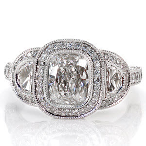 This tantalizing design puts all the focus on the 1.00 carat cushion cut center diamond. On either side of center is a half-moon diamond in a micro pavé halo. The band is lavishly adorned with diamonds on the top and sides. The halo displays pockets of filigree and a butterfly shape for a picturesque side view.