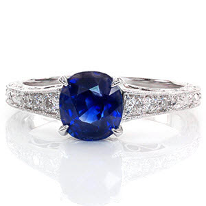 The lustrous blue of the sapphire center is decorated in a four prong claw setting for a pleasing top view. The three faces of the ring are adorned in an intricate hand engraved scroll pattern for a timeless statement. Graduating diamonds form the top of the band for brilliance and sparkle.  