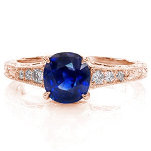 The lustrous blue of the sapphire center is decorated in a four prong claw setting for a pleasing top view. The three faces of the ring are adorned in an intricate hand engraved scroll pattern for a timeless statement. Graduating diamonds form the top of the band for brilliance and sparkle.  