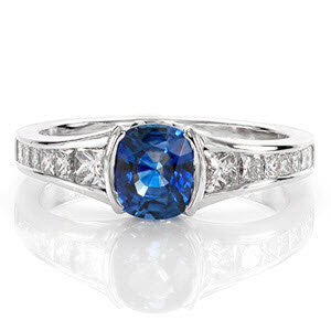 Design 1956 is a design of decisive lines and crisp form. A natural 1.00 carat cushion cut sapphire exhibits an impressive profile from a half bezel setting. Crafted in 14k white gold, the high polish band forms a slight flare towards the center stone with channel set graduated princess cut diamonds.