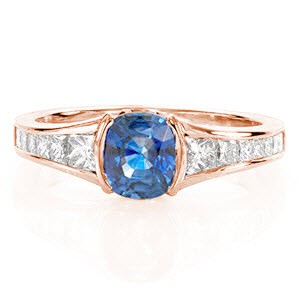 Design 1956 is a design of decisive lines and crisp form. A natural 1.00 carat cushion cut sapphire exhibits an impressive profile from a half bezel setting. Crafted in 14k white gold, the high polish band forms a slight flare towards the center stone with channel set graduated princess cut diamonds.