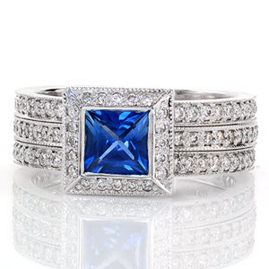 Magnificent lines of milgrain outline round bright-cut and bead-set micro pavé diamond bands that are joined into one distinctive wide band. Set atop the perfect square halo is a natural 0.75 princess cut blue sapphire. Crafted in 14k white gold, the full bezel captures the sapphire and completes a uniform design.