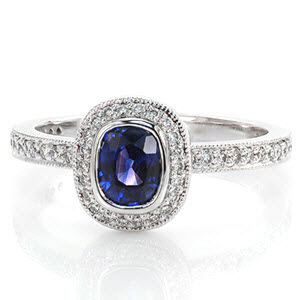 The soft curves of a regal 0.75 cushion cut blue sapphire shape the layout of Design 2048. In 14k white gold, the full bezel halo is surrounded by micro pavé diamonds. Formal lines of the micro pavé band create a pedestal for the halo, providing a striking profile and area for additional bands.