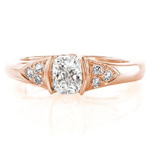 This charming design melds simple details to create a beautiful design. The 0.75 carat cushion cut center stone is held in a half-bezel setting and flanked on either side by a cluster of three round diamonds. The profile reveals graceful hand-formed filigree which adds a vintage feel to the ring. 