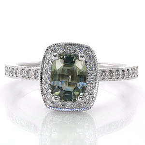 Design 2120 features a natural 1.25 carat mossy green cushion cut sapphire. The unique halo of micro pavé diamonds on the top and sides reveal filigree curls under the basket. The 14k white gold band arches up to join the halo for a sophisticated fit.