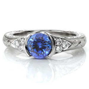 Sapphire Seville displays a 1.25 carat vibrant blue gemstone. The high polished half-bezel fashions the vivid round sapphire. A cluster of three diamonds flank each side the center stone. Hand engraved patterns and milgrain adorn this antique inspired ring for a picturesque view.