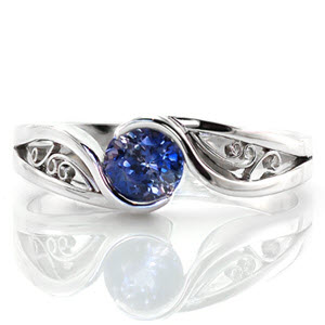 This mesmerizing solitaire design focuses on the luscious 0.80 carat round cut blue sapphire center. The band is a unique split shank with lots of movement and the ends flow around the center stone to form a half bezel setting. The pockets of the band are filled with graceful filigree curls.