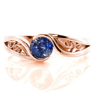 This mesmerizing solitaire design focuses on the luscious 0.80 carat round cut blue sapphire center. The band is a unique split shank with lots of movement and the ends flow around the center stone to form a half bezel setting. The pockets of the band are filled with graceful filigree curls.
