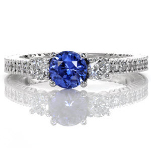 Modern charm meets a timeless three stone classic in this contemporary engagement ring. The deep blue 0.80 carat round sapphire is intensified between the two round brilliant diamonds. Micro pavé stones heighten the beauty of the textured band for a unified look. 
