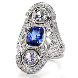 This vintage inspired wide ring design features a 1.00 carat royal blue sapphire. The vertical design also features two large full bezel set diamonds. The ornate pattern is decorated with pierced elements that are embellished in micro pavé set diamonds. This design is truly a statement piece.