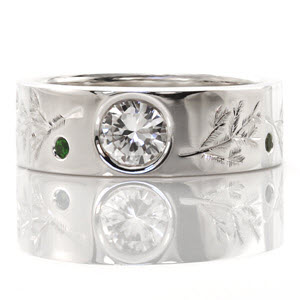 This custom design features unique engraved designs of pine branches. The center stone is a bezel set 0.80 carat round brilliant cut diamond. The wide band also has vivid green emeralds set in between the engraving. The wide band adds a contemporary touch to the ring.