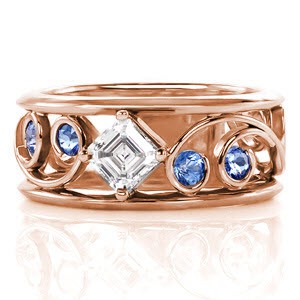 Classic filigree is paired with this modern wide band for stylish appeal. Each intricate curl is adorned with a Ceylon sapphire for a hint of color. Interwoven between the scroll patterns is a 0.50 carat asscher cut diamond. The linear parallel facets give this center stone the appearance that it goes on forever.    