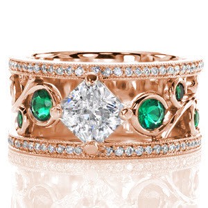 Vivid green emerald side stones are bezel set into the large scroll of the filigree pattern. The 1.00 carat radiant cut center diamond is kite-set to continue the flow of the filigree. A row of micro pavé adorns both sides of this wide band ring with milgrain texture to accent the edges. 
