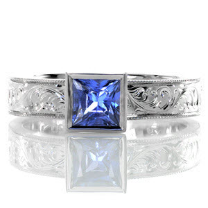 The sumptuous pop of bright blue from the 0.75 carat princess cut sapphire makes this design simply exquisite. The bezel set stone is accented by a wide band with fascinating hand engraved designs which are intricately carved one line at a time. The edges are given a refined finish with milgrain.