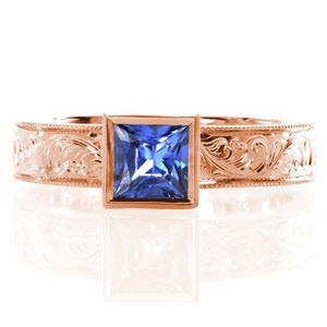 The sumptuous pop of bright blue from the 0.75 carat princess cut sapphire makes this design simply exquisite. The bezel set stone is accented by a wide band with fascinating hand engraved designs which are intricately carved one line at a time. The edges are given a refined finish with milgrain.