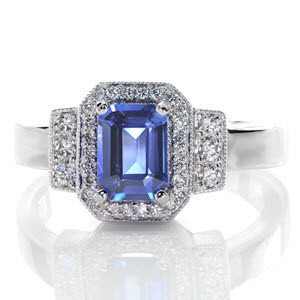 A luxurious deep blue 1.00 carat emerald cut sapphire is the center of attention in this Art Deco inspired piece. The stone is contrasted with bright white micro pavé diamonds set within a halo, as well as in the rectangular bead set shoulder detail. The remaining elements are left high polished.
