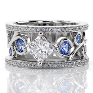 This stunning wide band has a radiant pop of color from the blue sapphires set into the curls of the large filigree. The center stone is a kite-set 1.00 carat princess cut diamond that continues the flow of the filigree design. The band is completed with two rows of micro pavé and milgrain edging.