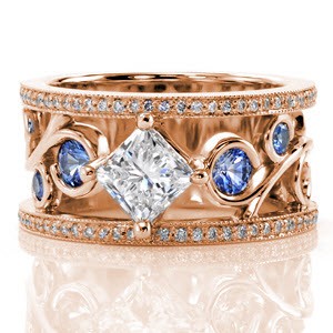This stunning wide band has a radiant pop of color from the blue sapphires set into the curls of the large filigree. The center stone is a kite-set 1.00 carat princess cut diamond that continues the flow of the filigree design. The band is completed with two rows of micro pavé and milgrain edging.