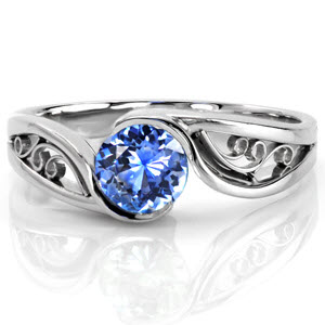 Design 2385 is a sophisticated solitaire featuring a natural 0.75 carat round cut blue sapphire. The high polish band portrays flowing movement as it embraces the center stone with a half bezel. The open pockets between the split shank make the perfect frame for filigree curls, keeping the design delicate.