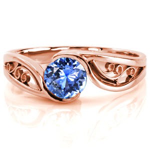 Design 2385 is a sophisticated solitaire featuring a natural 0.75 carat round cut blue sapphire. The high polish band portrays flowing movement as it embraces the center stone with a half bezel. The open pockets between the split shank make the perfect frame for filigree curls, keeping the design delicate.