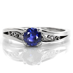 A 0.60 carat round cut natural blue sapphire is set in a classic four prong setting. Repeating the curves of the center stone, rhythmic scrolls of hand wrought filigree decorate the pockets created by the split shank. Crafted in 14k white gold, the bands unique split creates a soft waved contour.