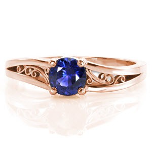A 0.60 carat round cut natural blue sapphire is set in a classic four prong setting. Repeating the curves of the center stone, rhythmic scrolls of hand wrought filigree decorate the pockets created by the split shank. Crafted in 14k white gold, the bands unique split creates a soft waved contour.