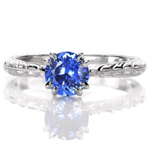 The rich, vibrant blue against the bright, white luster of the metal is a breathtaking sight. A 1.0 carat round blue sapphire is securely held by claw prongs and is detailed at the bottom with two surprise diamonds. The milgrain knife-edge is accented with wheat pattern engraving on each side. 