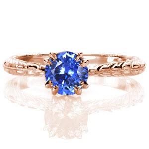 The rich, vibrant blue against the bright, white luster of the metal is a breathtaking sight. A 1.0 carat round blue sapphire is securely held by claw prongs and is detailed at the bottom with two surprise diamonds. The milgrain knife-edge is accented with wheat pattern engraving on each side. 