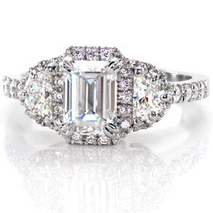 This halo engagement ring features a 1 carat emerald cut and two half moon shaped diamonds. The center stone is prong set with a double prong on each corner. Around these three stones is a halo of small micro pave set diamonds. Both sides of this design feature hand wrought filigree in three pockets.