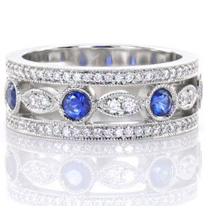 The outside rows of the Sappire Dolce are adorned with micro pavé diamonds and milgrain detail. Alternating marquise shaped patterns and circular outlines detail the center of the design. Vivid blue sapphires fill the circular arrangement and two round brilliant diamonds accent the marquise shaped silhouettes.