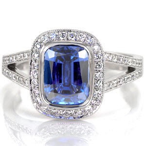 The royal blue of the 2.00 carat cushion cut sapphire is a signature statement of regal expression. Round diamonds outline the sapphire to embellish the beautiful blue of the center stone. The split shank cathedral is lined with bead-set diamonds and accented with two bezel set stones for a majestic finish.  