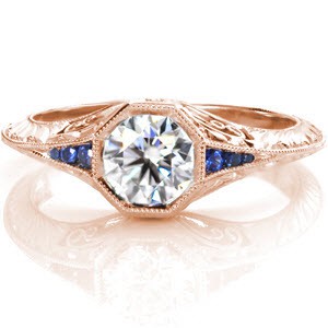 This stunning antique inspired ring features a round brilliant cut center stone in an octagonal setting with a knife-edged band. The luscious blue sapphire side stones are captivating and keep the eye moving around the whole piece. The band is further detailed with exquisite hand engraving and hand made filigree curls.