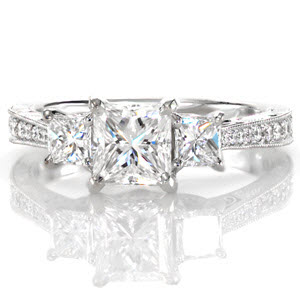 This stunning princess cut three stone setting is elegantly accented with a row of round brilliant diamonds along the length of the band. Intricate antique inspired details of milgrain, filigree and hand engraving embellish the side face of this design. Five petals are embellished with diamonds for a radiant appeal.  