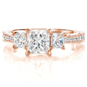This stunning princess cut three stone setting is elegantly accented with a row of round brilliant diamonds along the length of the band. Intricate antique inspired details of milgrain, filigree and hand engraving embellish the side face of this design. Five petals are embellished with diamonds for a radiant appeal.  