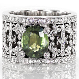 This beautiful custom engagement ring features a 2.50 carat oval cut green sapphire center. The rich, earthy green is highlighted by the intricate curls of white gold that form the pattern of the band. The curls of the filigree pattern, and of the center prongs, are bezel set with brilliant diamonds.