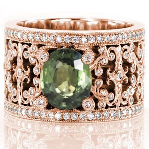 This beautiful custom engagement ring features a 2.50 carat oval cut green sapphire center. The rich, earthy green is highlighted by the intricate curls of white gold that form the pattern of the band. The curls of the filigree pattern, and of the center prongs, are bezel set with brilliant diamonds.