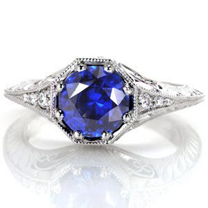 Design 2855 is a stunning example of elegance with a rich blue hue of the 1.25 carat round cut sapphire taking center attention. Crafted in 14k white gold, single wheat hand engraving and graduating round cut diamonds adorn the knife edge band. Surprise stones and hand formed filigree curls accentuate a vintage look.