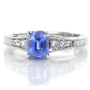 A vibrant blue cushion cut sapphire center stands out in this custom, antique inspired engagement ring. The band flares as it reaches the center and is adorned with round diamonds that graduate in size. The top and sides of the band are detailed with hand engraved scroll designs, and hand formed filigree curls.