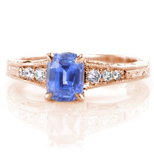 A vibrant blue cushion cut sapphire center stands out in this custom, antique inspired engagement ring. The band flares as it reaches the center and is adorned with round diamonds that graduate in size. The top and sides of the band are detailed with hand engraved scroll designs, and hand formed filigree curls.