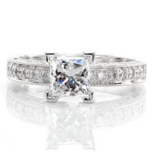 The Adorable Princess is a darling design with a 1.00 carat princess cut diamond. The top of the band is accented by large, bead-set stones. The sides feature bezel set diamonds and cut outs, including a heart shape on the side of the center stone. Milgrain detail adds texture to the edges.