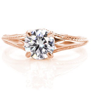This custom solitaire engagement ring features a round brilliant cut diamond displayed in a knife edge band. The detailed hand engraving displays intricate wheat and scroll patterns for a vintage appeal. The hand formed filigree in the basket of the center stone setting creates a focal for this antique inspired design.