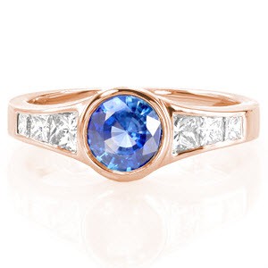 The 1.00 carat vivid blue sapphire is fashioned in a white gold bezel putting emphasis on the cobalt hue of the center stone. Princess cut side stones graduate in size drawing the eye to the beautiful sapphire center. The high polished metal gives this contemporary design a very sleek appearance. 
