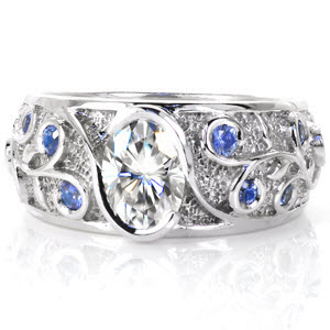 Stunning wide filigree engagement ring set with pale blue sapphires in Denver. The oval cut diamond center stone is held in a bezel setting, with blue sapphires bezel set in the filigree curls. Filigree forms leaf and vine patterns on a stippled background. 