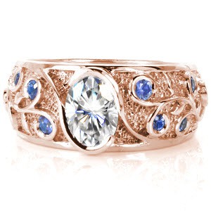 This custom wide band features an oval cut diamond center in a half-bezel wrap setting. The elegant flow of the relief-engraved filigree pattern compliments the center setting. The stippled background highlights the polished luster of the vines, while blue ceylon sapphires sparkle from the curls. 