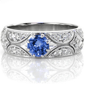 This glamorous design features a 0.75 carat round sapphire in an eight prong setting. The band is set with crescents of micro pavé and star burst patterns to create a glittering display of light. Beaded milgrain texture outlines each section for added definition. 