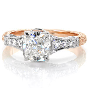 The stunning cushion cut center is fashioned in this exquisite antique inspired setting. The shine of the white metal is decorated with brilliantly faceted diamonds while the warmth of the rose gold is adorned with relief hand engraving. Milgrain texture and platinum filigree enhances its vintage appeal.    