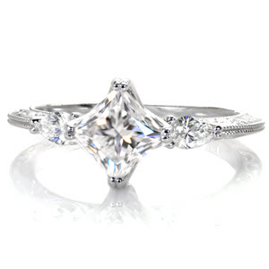 This elegant three stone diamond engagement ring is breathtaking with a kite-set princess cut center diamond. The antique inspired knife edge band flows beautifully down from the points of the pear cut side stones. This unique engagement ring is exquisitely detailed with relief-style hand engraving and milgrain.
