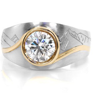 This custom design is crafted in two-tone platinum and 18K yellow gold.  It features picturesque hand engraving of a mountainous skyline framing the central gemstone.  A 1.00 carat round brilliant diamond is accented by a curving full bezel in a contrasting vivid yellow gold.   