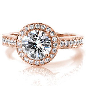 A 1.20 carat round brilliant cut center is set in four prongs and surrounded by a halo of micro pavé stones. Diamond bands join under the halo and are accented with surprise diamonds to add a delightful charm to the side-profile of this design. The band is detailed stones and milgrain to match the diamond halo.  